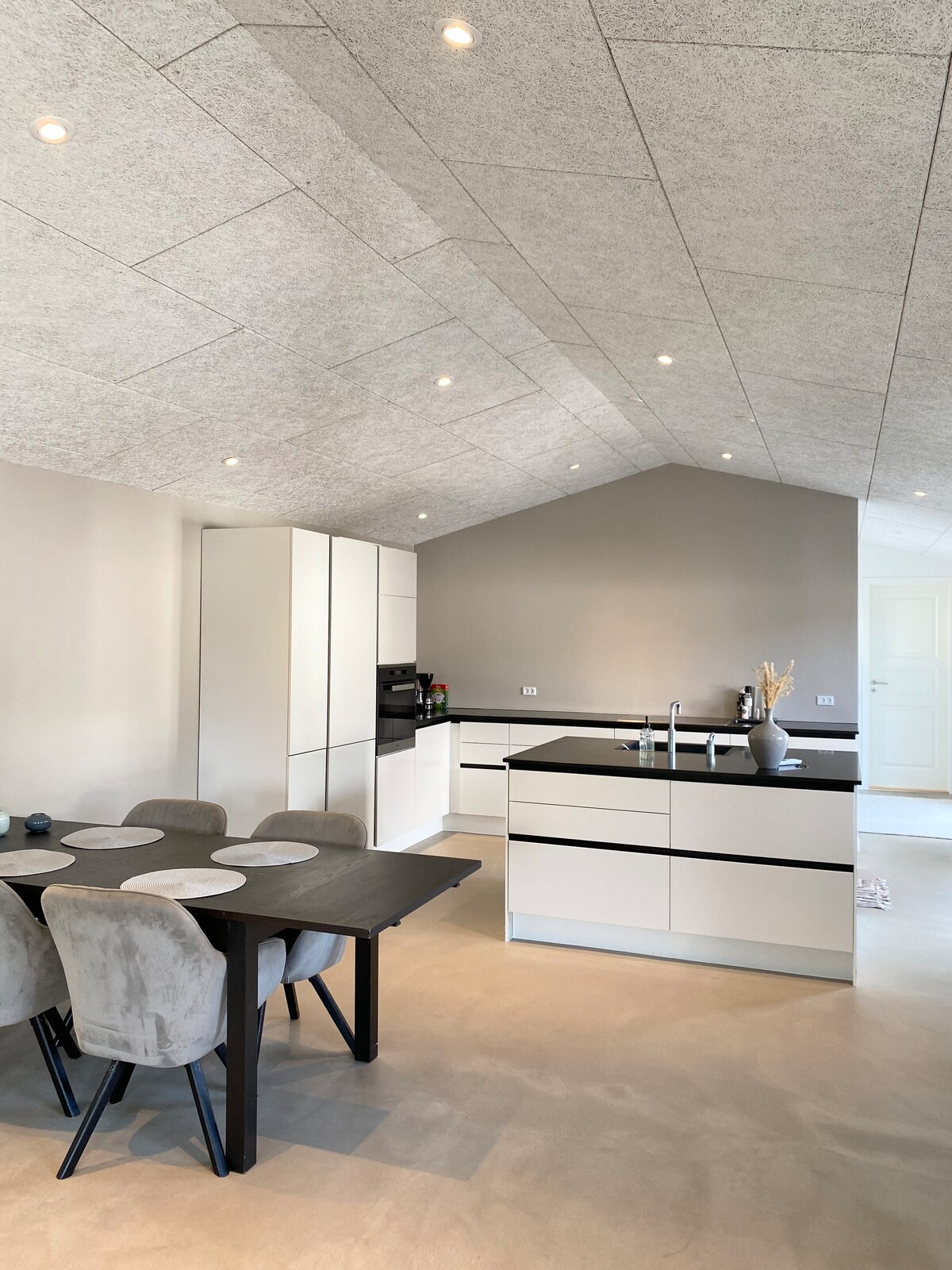 A modern kitchen area features sleek white cabinetry contrasted with a dark countertop. An island with seating is positioned at the center, surrounded by a simple dining table and chairs. Recessed lighting enhances the spacious, airy atmosphere presented by the high, sloped ceiling.