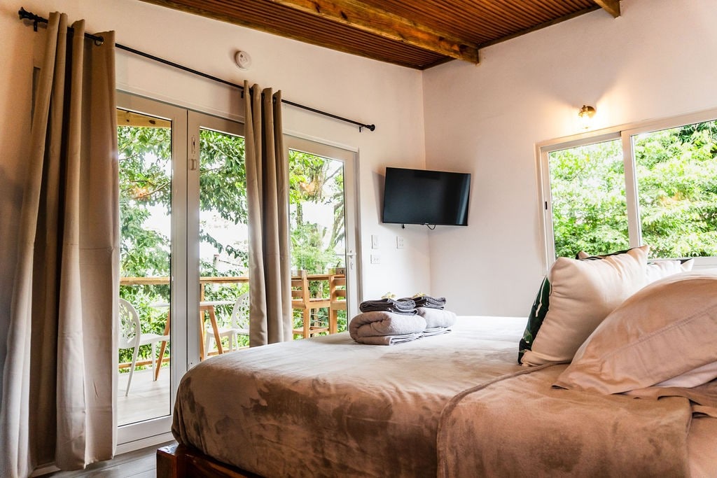 A spacious bedroom features a large bed covered with soft linens. A wall-mounted TV is positioned nearby, while large windows provide a view of lush greenery and allow natural light to fill the room. A private balcony is accessible from the sleeping area.