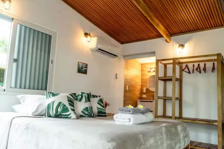 Chambre lumineuse et climatisée avec un lit confortable, des serviettes pliées de qualité, des oreillers moelleux et un placard en bois. La salle de bain privée en arrière-plan met en valeur le design soigné et le confort de cette cabane dans la jungle de Big Creek à Bocas del Toro.