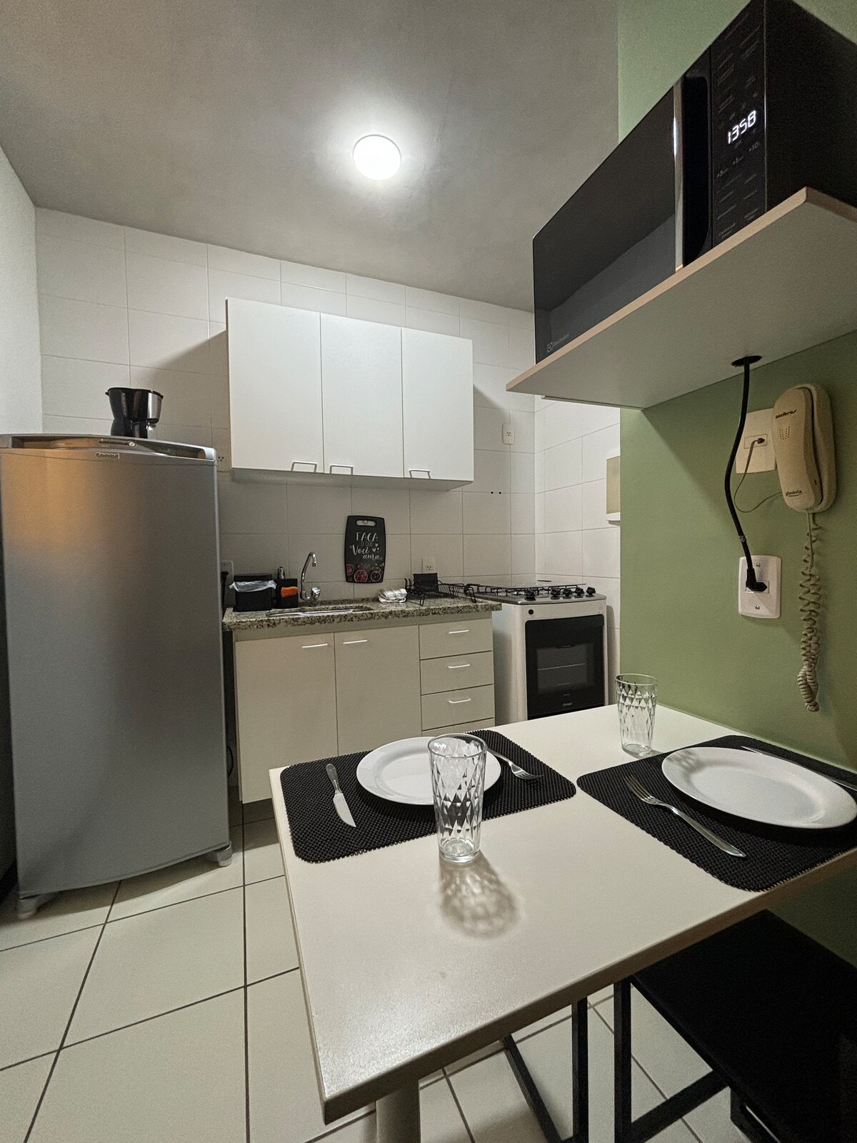 A compact kitchen area features light-colored cabinetry and a stainless-steel refrigerator. A gas stove is positioned alongside a microwave on a shelf. A small dining table is set for two, accompanied by clear glasses and white plates, creating a practical and functional space.