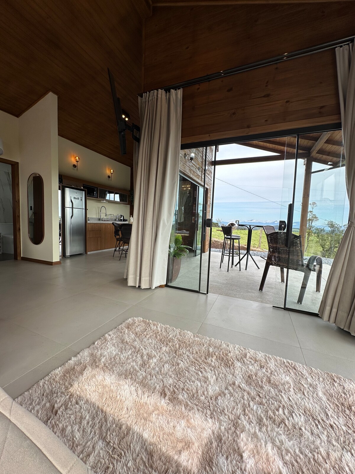 An open living area features a plush area rug in the foreground, leading to large glass doors that provide a view of the outdoor seating. A modern kitchen is visible in the background, complemented by warm wood accents and natural light streaming into the space.