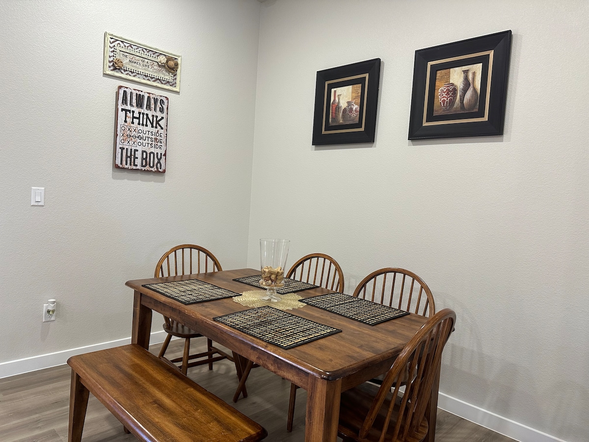 A wooden dining table accompanied by six chairs is positioned against a light-colored wall. Two decorative frames hang above the table, while a centerpiece with a glass vase holds small decorative items. Textured place mats are laid out on the table surface.