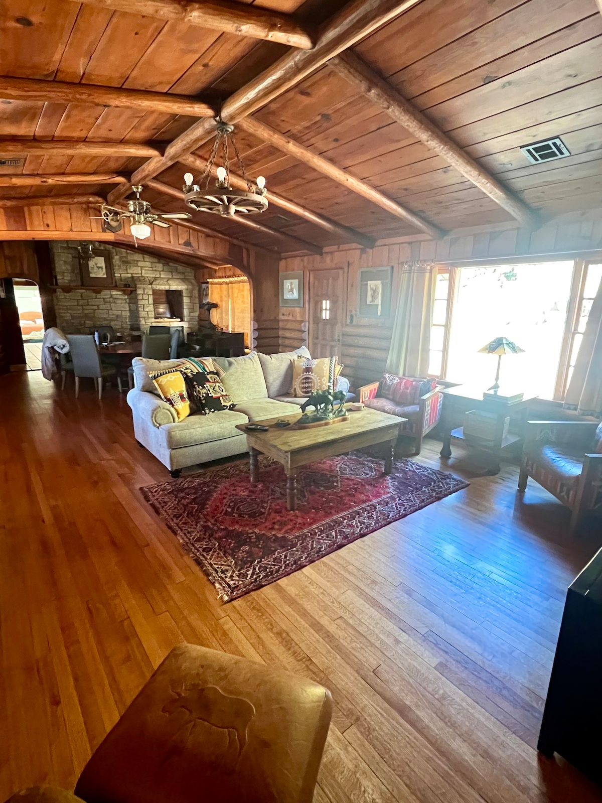 A cozy living area features rustic wooden beams overhead, with a comfortable sofa adorned with vibrant cushions. A wooden coffee table sits on a patterned area rug, surrounded by armchairs and a small lamp, while large windows provide natural light.