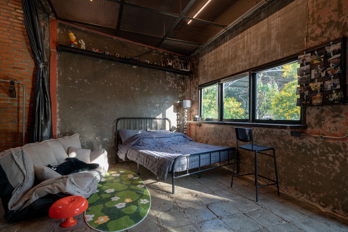 A spacious loft-style bedroom features a queen-size bed, complemented by a cozy sofa and a black chair. Natural light filters through large windows, illuminating the textured walls. A round area rug adds a touch of color to the industrial design.