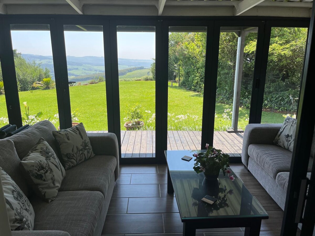 A sitting area is oriented towards expansive glass doors that open to a grassy landscape and distant hills. The space features a light-colored sofa with decorative cushions and a dark coffee table. Natural light fills the room, creating a bright and airy environment.