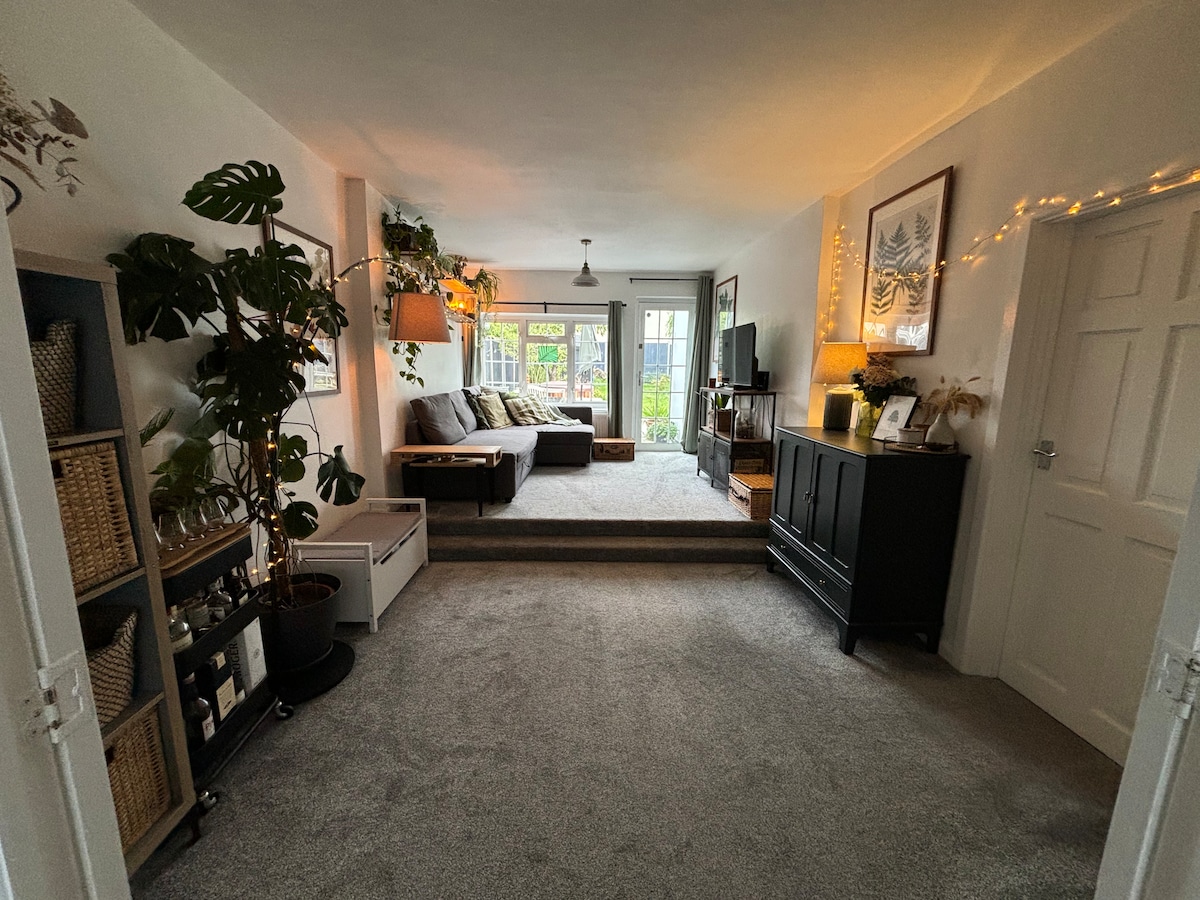 The spacious living room features soft grey carpeting and large windows that allow natural light to fill the space. A comfortable sectional sofa is arranged on a raised area, with a TV and decorative plants adding to the inviting setup. Accessible storage options are visible along one wall.