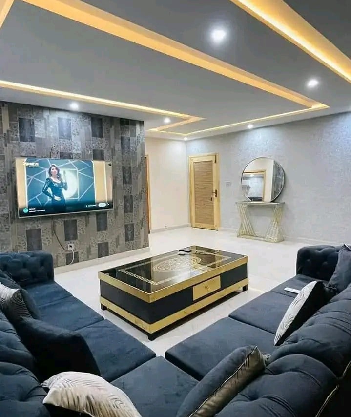 A modern living room features a dark blue sectional sofa arranged around a sleek coffee table with decorative design. A large flat-screen TV is mounted on the wall, while a round mirror and a wooden cabinet are positioned nearby. Soft ambient lighting enhances the space.