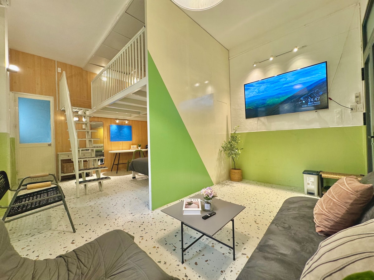 A cozy living area features a comfortable sofa and a black coffee table, with a small plant adding a touch of greenery. A large TV is mounted on the wall, and a white staircase leads to the lofted bed area. Soft natural light enters through the windows.