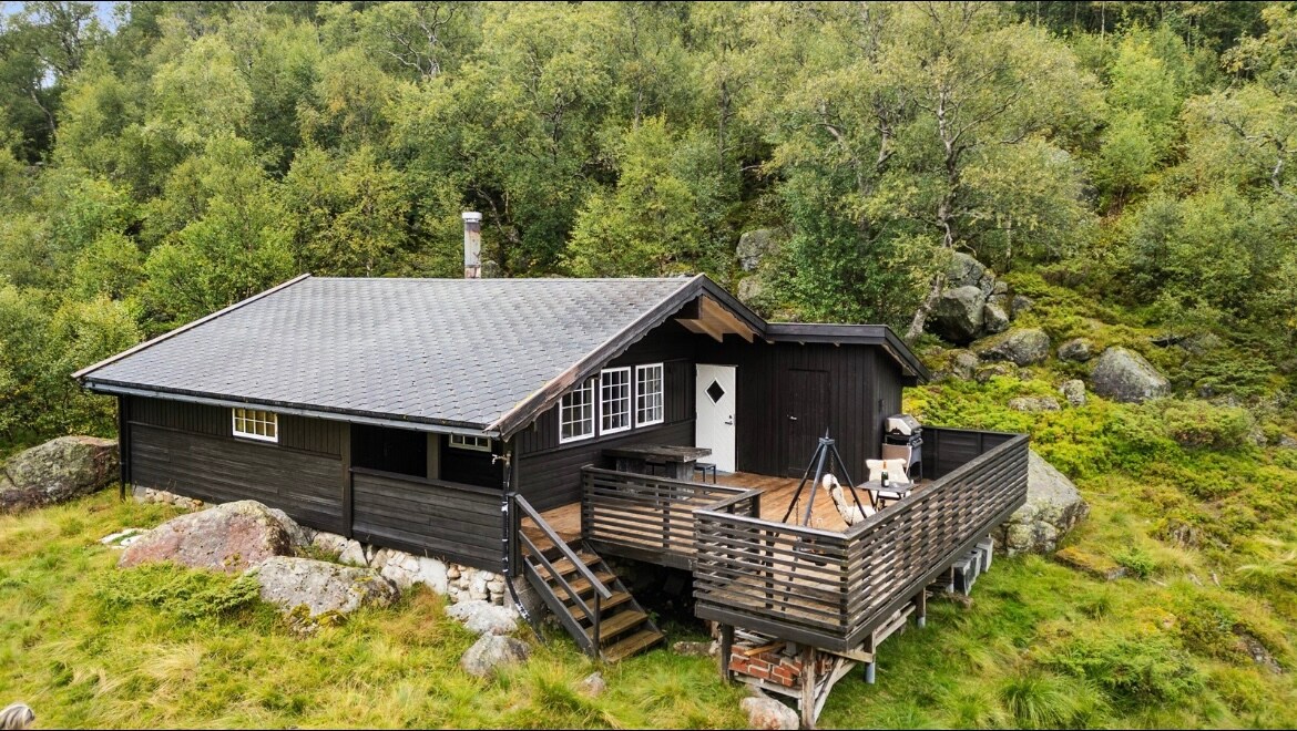 A rustic wooden cabin is set amidst lush greenery, featuring a spacious deck with wooden railings. The sloping roof and chimney are visible, complemented by a stone foundation. Large windows allow natural light, while steps lead to the entrance, surrounded by a serene landscape.