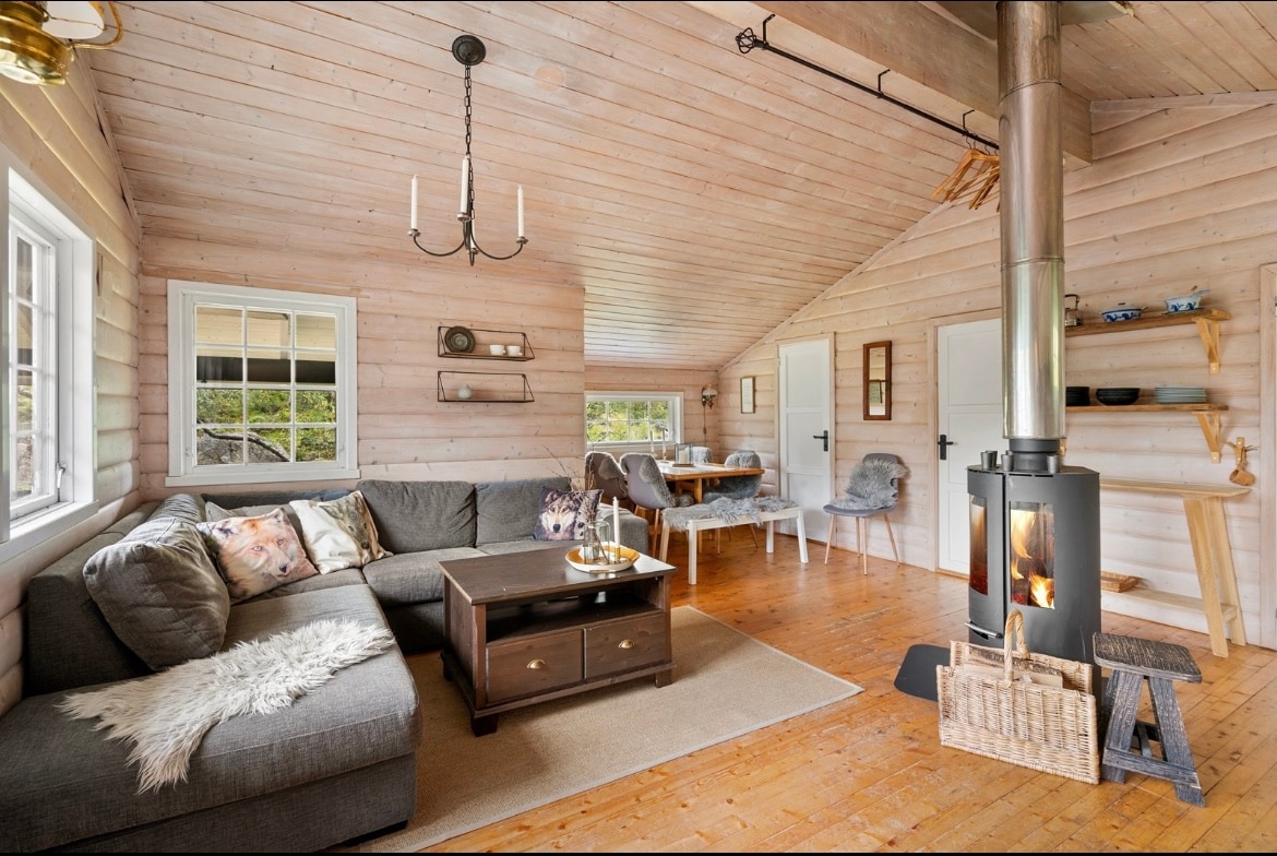An inviting living area features a comfortable gray sofa adorned with decorative pillows, coupled with a wooden coffee table. Natural light floods through large windows, illuminating the warm wooden walls. A dining table with chairs is visible in the distance, beside a modern wood stove providing warmth.