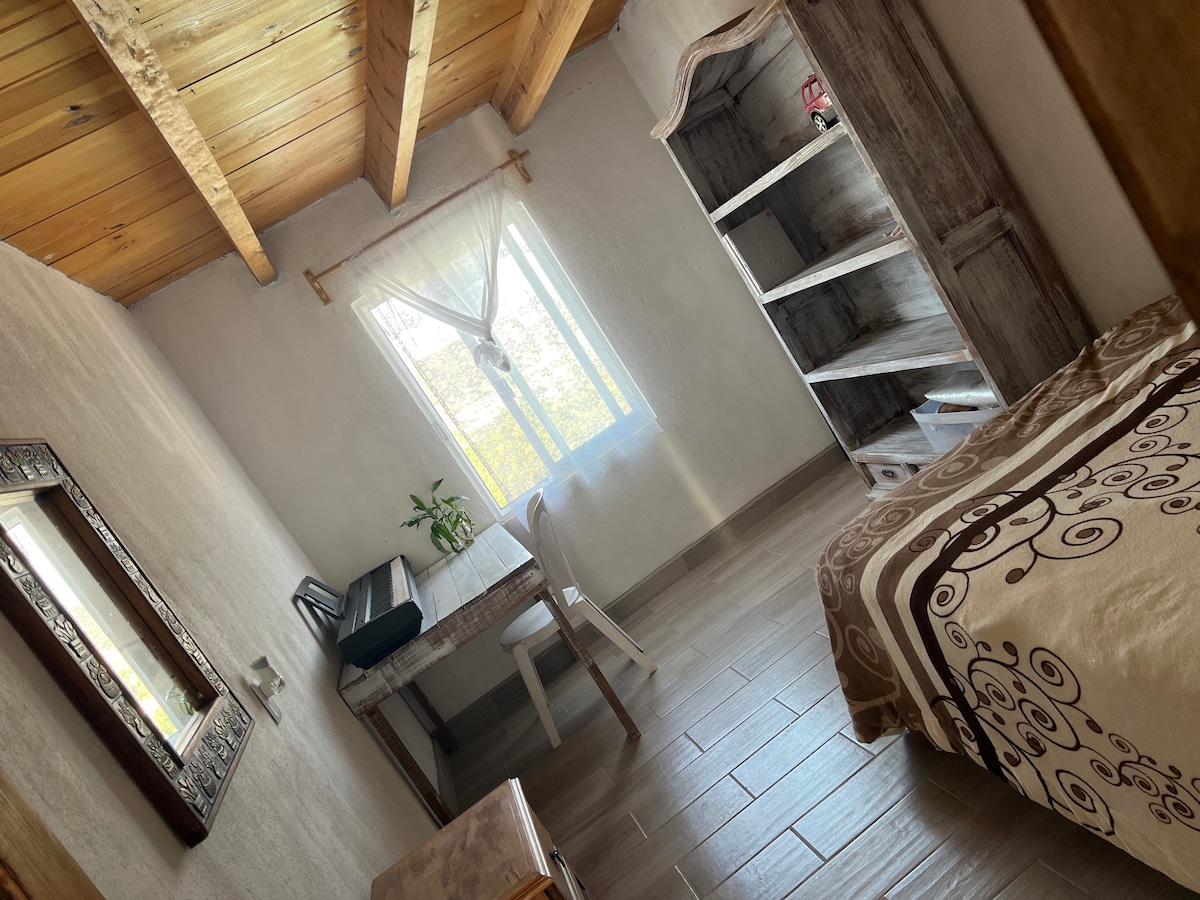 A serene bedroom features a single bed with a brown and white patterned coverlet. Natural light enters the room through a window with sheer curtains. A wooden desk and chair are positioned beside the window, with a shelf displaying decor items in the corner.