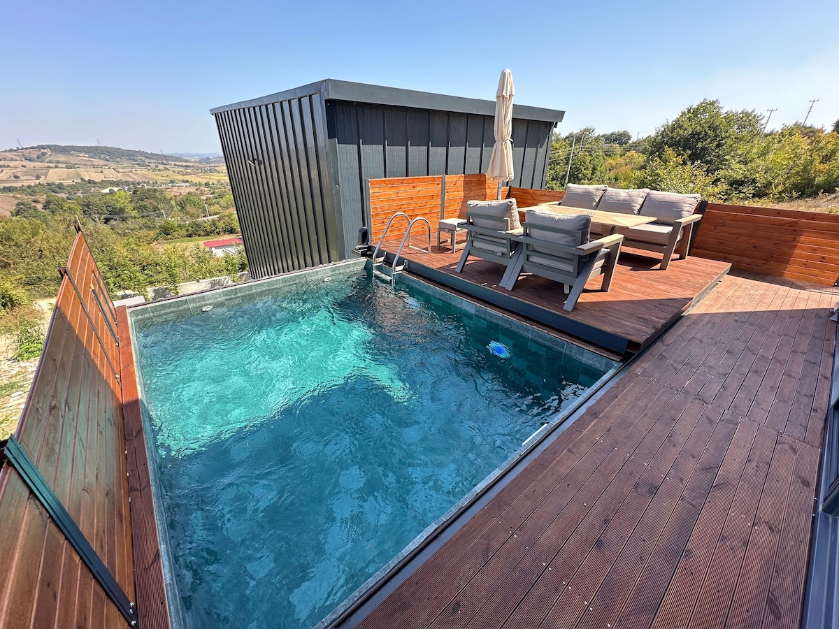 An outdoor space features a private swimming pool surrounded by wooden decking. Comfortable seating arrangements are positioned nearby, accommodating relaxation. The backdrop showcases a rural landscape, providing a serene setting under clear blue skies.