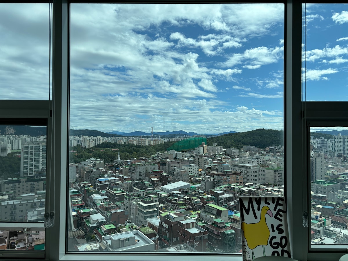 An expansive window reveals a panoramic view of a city landscape, featuring densely packed buildings and green hills in the distance. A colorful artwork sits on the windowsill, adding a touch of vibrancy to the scene. The sky is partly cloudy, enhancing the natural light.