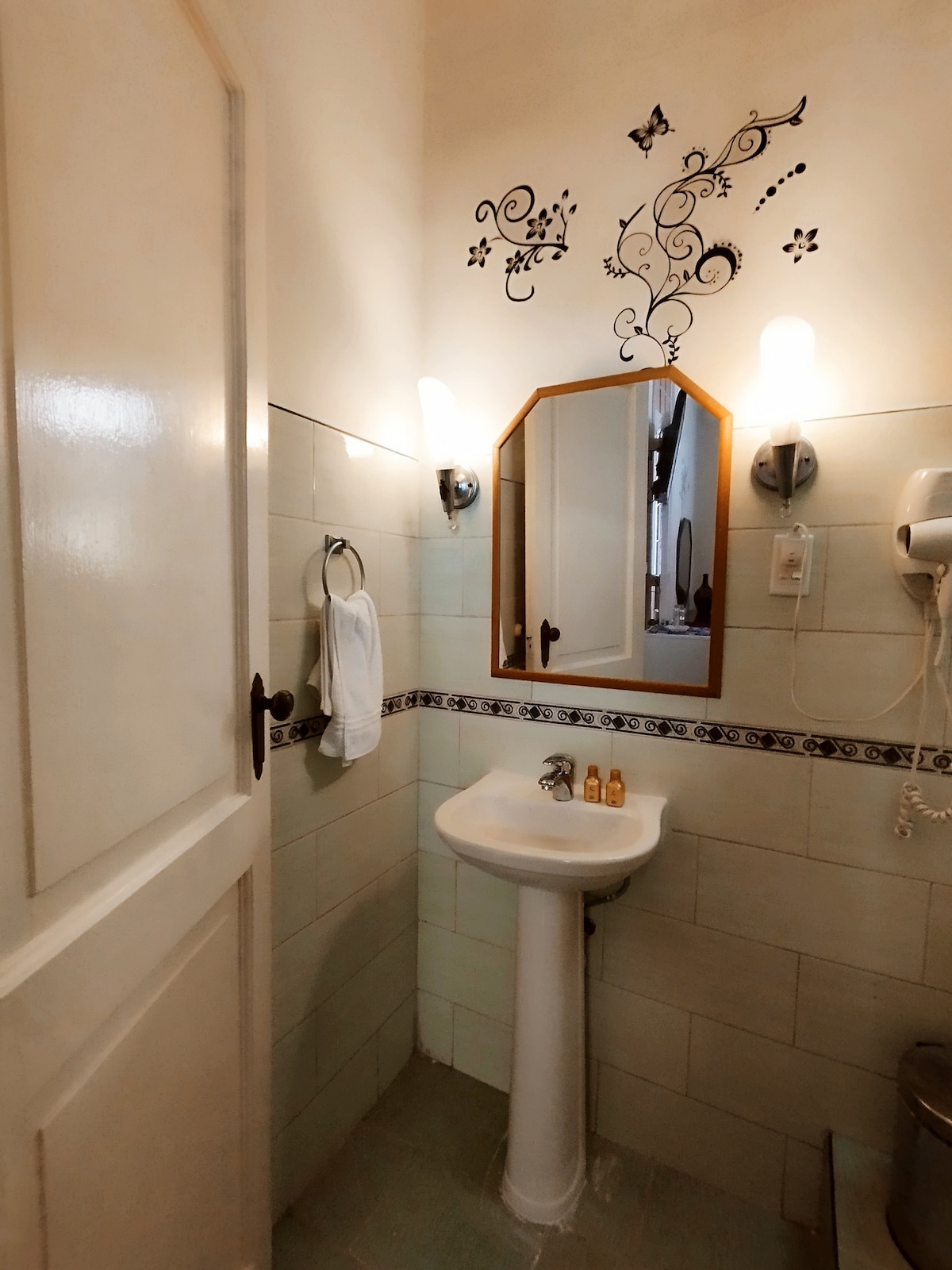 A compact bathroom features light-colored tiles and a small pedestal sink, complemented by a decorative mirror. Wall-mounted light fixtures provide illumination. A towel hangs neatly on a hook, and a hairdryer is attached nearby, enhancing the bathroom's functional design.