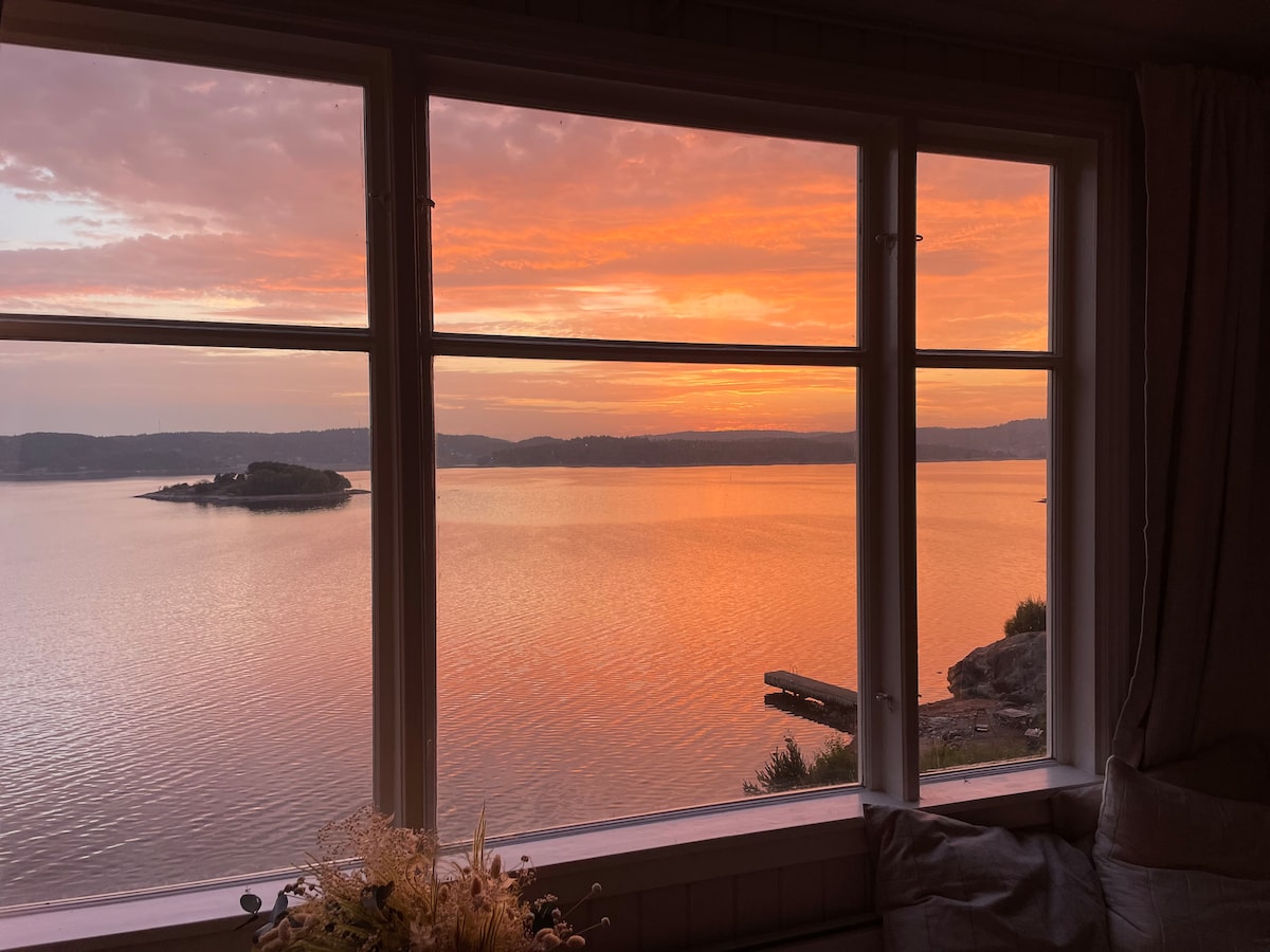A scenic view of a sunset is framed by large windows, showcasing vibrant colors reflecting off calm water. An island is visible in the distance, and a boat dock can be seen nearby. Soft light illuminates the interior, creating a serene ambiance.