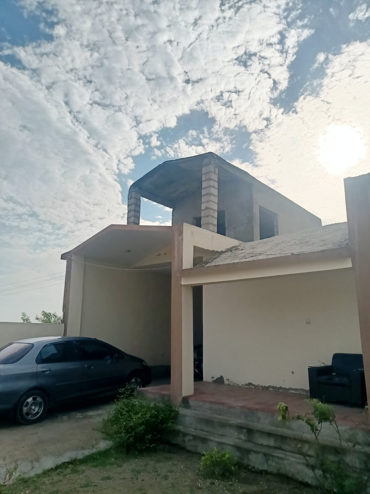 A modern two-story house is shown, showcasing a stone and concrete facade under a partly cloudy sky. A gray car is parked on the paved surface in front of the house. The entrance features a covered porch that leads into the inviting space.