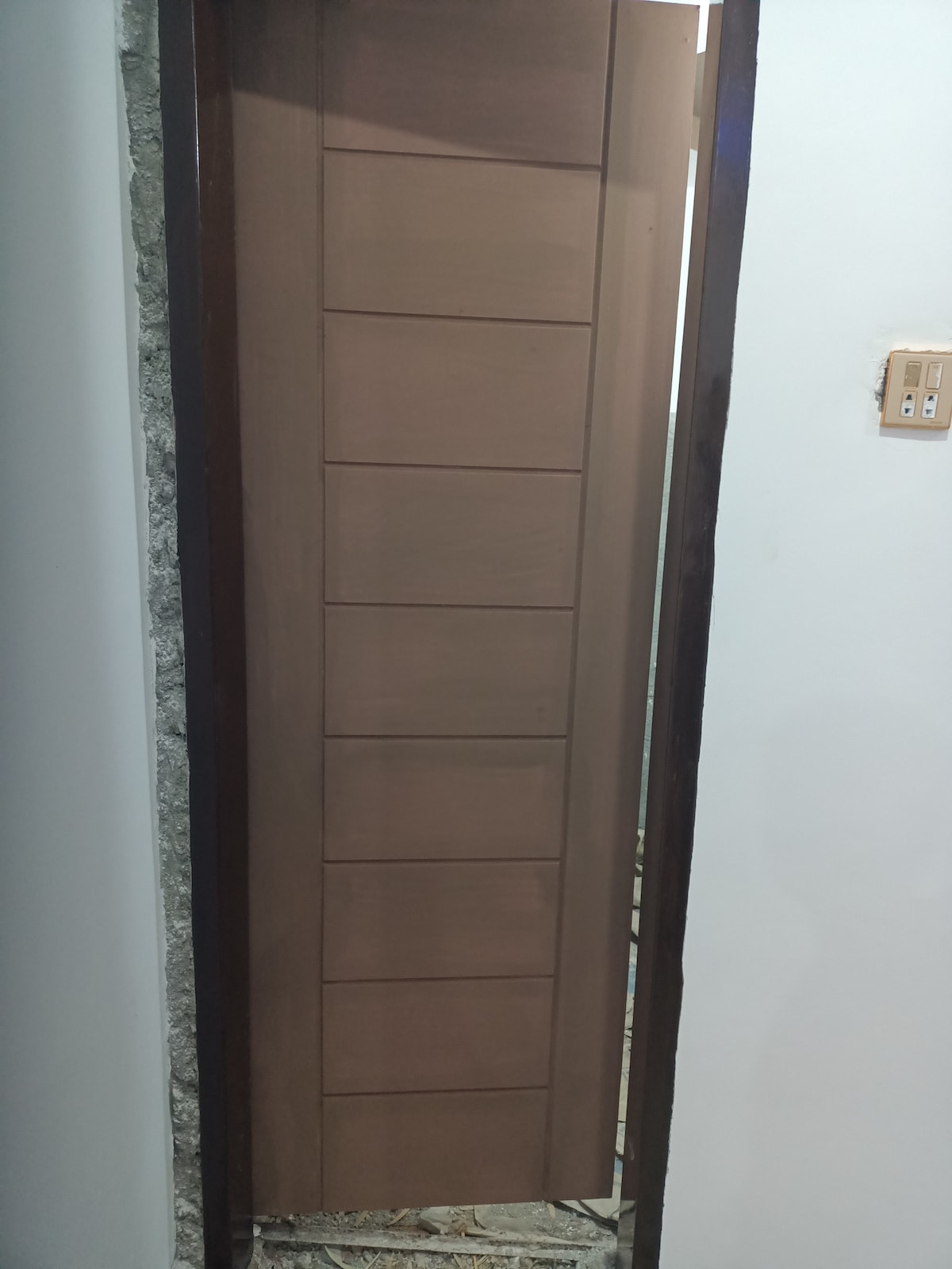 A simple wooden door is shown, featuring a paneled design. The door is partially ajar, revealing hints of another space beyond. The surrounding walls are painted in a neutral tone, with a stone accent visible alongside the doorway.