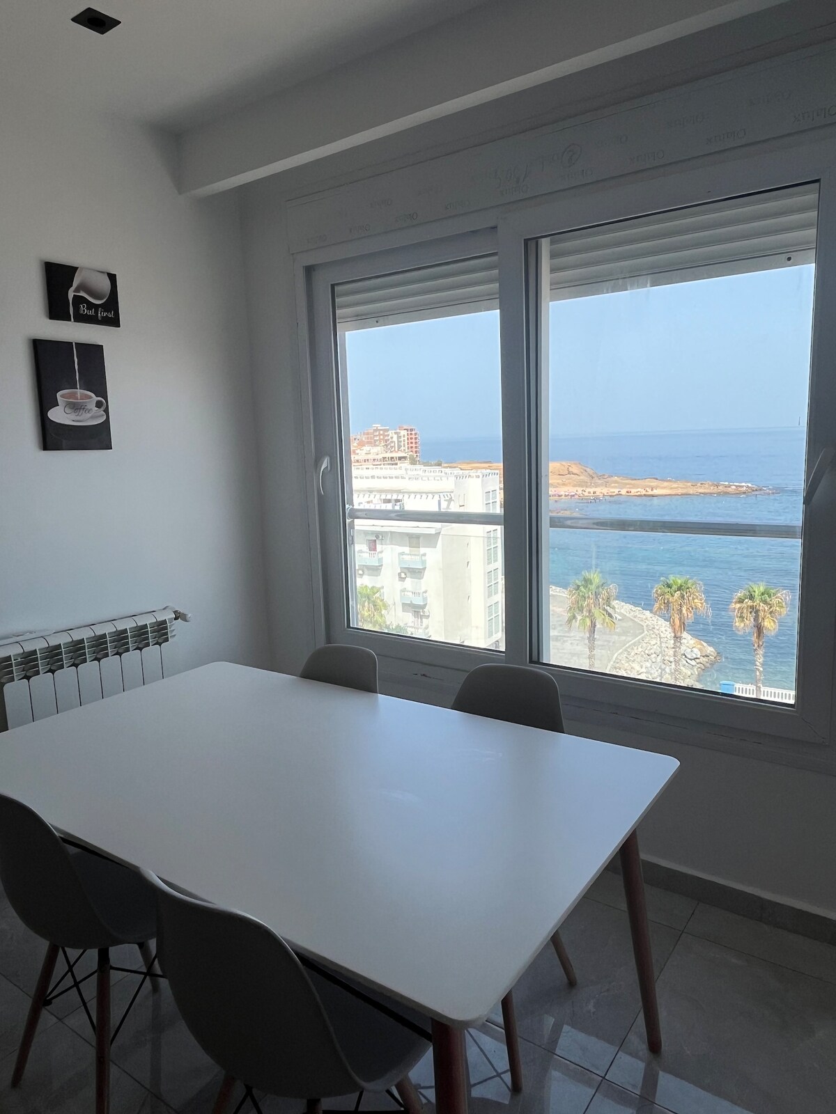 A dining area features a white table surrounded by four chairs, positioned near a large window with stunning sea views. The bright space is enhanced by natural light, while framed art pieces adorn the wall, adding a touch of decor.