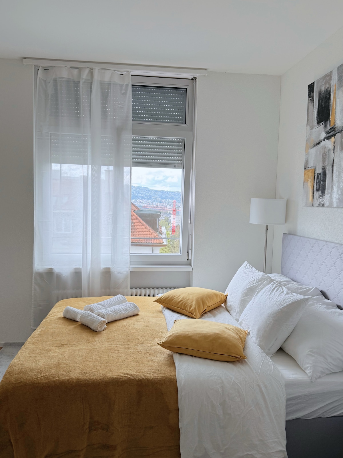 A bright bedroom features a spacious bed layered with white linens and a soft mustard throw. A pair of white towels is neatly arranged at the foot of the bed. Sheer curtains frame a window that overlooks a scenic view, allowing natural light to fill the room.