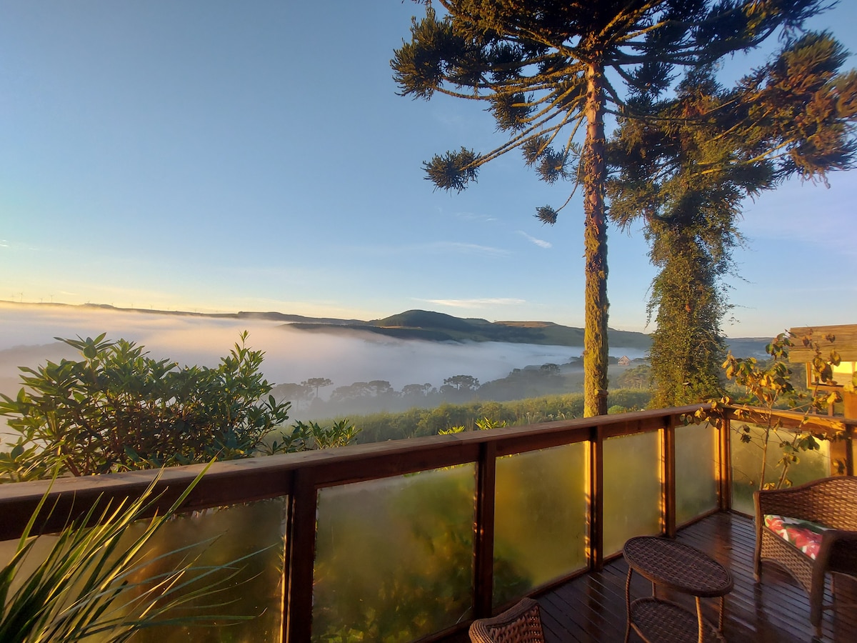 A panoramic view from the balcony showcases misty valleys and rolling hills under a clear sky. Lush greenery frames the scene, while sunlight filters through foliage, creating a serene atmosphere. A wooden railing borders the balcony, complementing the surrounding natural beauty.