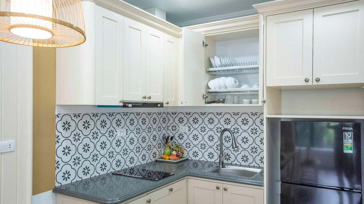 A clean and functional kitchen space features white cabinetry and an intricate patterned backsplash. A stainless steel refrigerator is positioned adjacent to a sink and stovetop. Open shelves display neatly arranged dishes, while a fresh bowl of fruit adds a touch of color to the countertop.