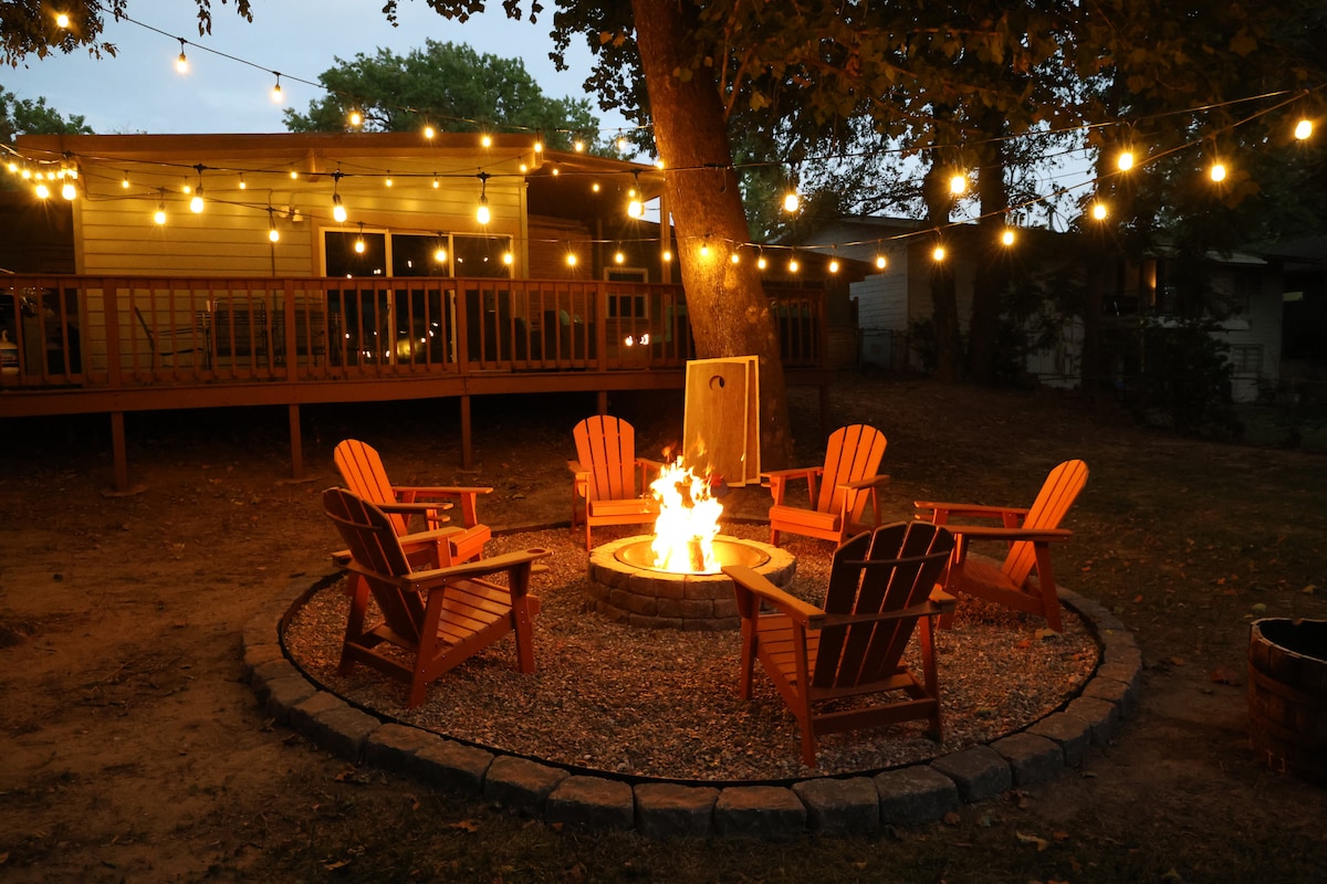 An outdoor fire pit area is surrounded by five adirondack chairs, inviting relaxation during evenings. Twinkling string lights are strung above, illuminating the space beneath a canopy of trees. The warm glow of the fire enhances the cozy atmosphere, highlighting the deck of the home in the background.