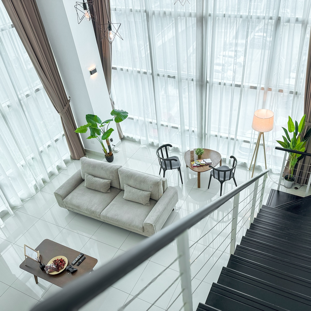 A light-filled living area features a contemporary sofa facing large windows with sheer drapes. A small dining table and chairs are positioned nearby, and a tall floor lamp provides ambient light. Potted plants add a touch of greenery to the minimalist decor.