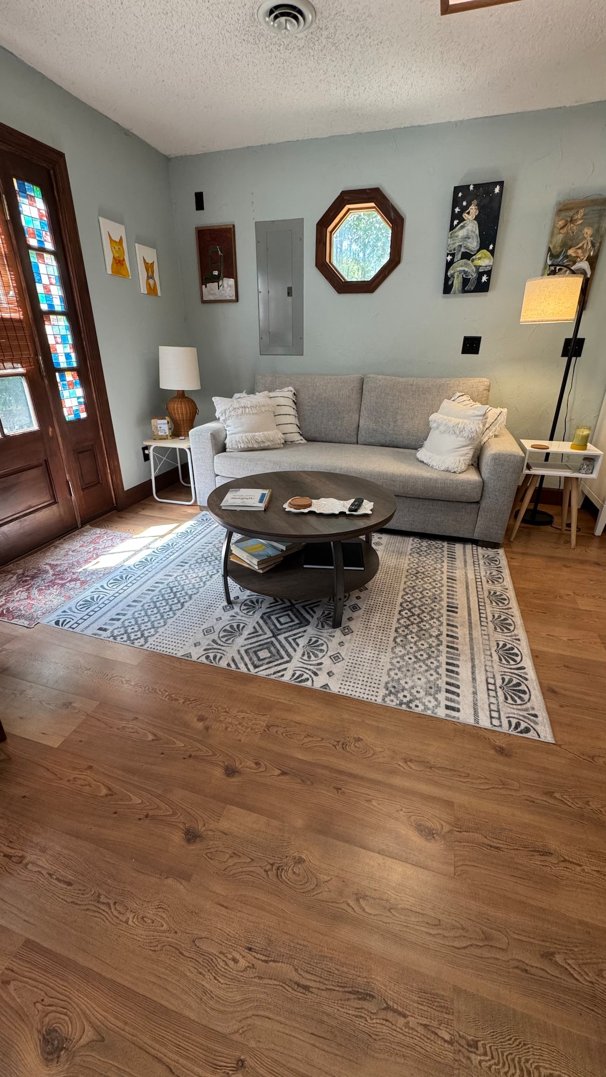 A cozy living area features a light gray couch with decorative pillows, positioned across from a circular coffee table. Natural light enters through a nearby window, illuminating the wood flooring and a patterned area rug. Colorful artwork adorns the walls, creating a welcoming space.