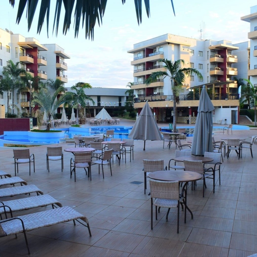 An outdoor area is depicted, featuring several lounge chairs and tables arranged around a modern pool. Umbrellas provide shade, and palm trees enhance the tropical ambiance. The buildings in the background house various facilities, creating a resort-like atmosphere.