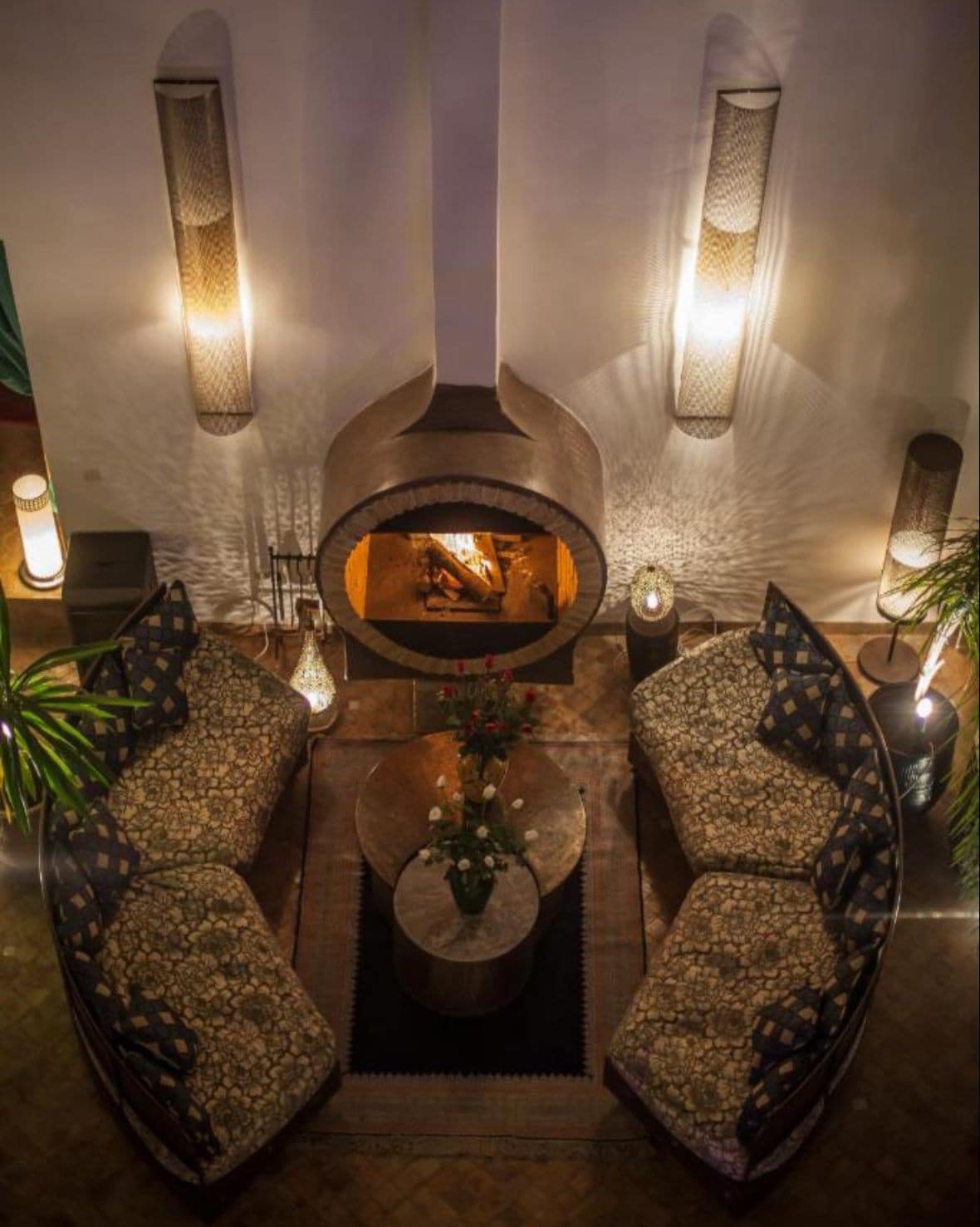 A cozy sitting area features curved seating upholstered in patterned fabric, arranged around a circular wooden table. A centrally located fireplace provides warmth and is framed by lush greenery, while wall-mounted lamps create soft, ambient lighting throughout the space.