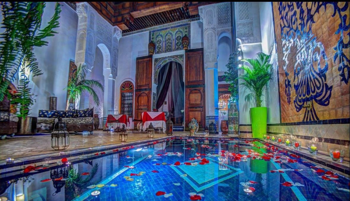 An elegant indoor space features a tranquil pool adorned with floating rose petals. Decorative lanterns and lush greenery complement the intricate tile work. Traditional seating areas are arranged beside the pool, creating a serene atmosphere for relaxation.