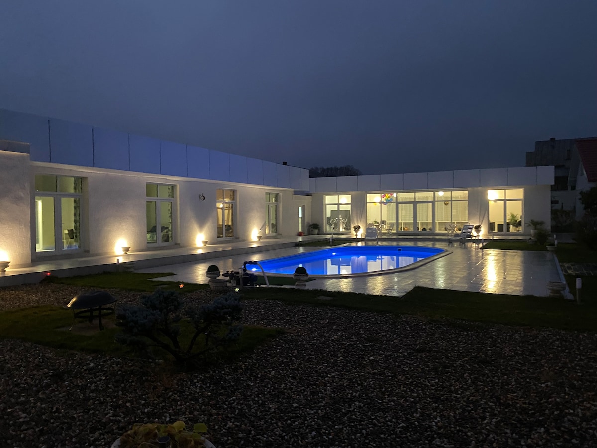 An evening view of a modern home featuring a well-lit outdoor area. A rectangular pool is situated in the center, reflecting soft blue hues. Surrounding pathways are illuminated by ground lights, and large windows provide glimpses of the interior space.