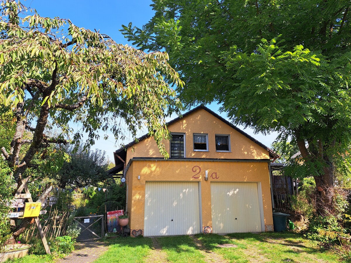 A charming yellow exterior of a two-story building is visible, featuring two garage doors and a welcoming entrance. Lush greenery surrounds the structure, with trees providing shade. Bright blue skies create a serene backdrop, enhancing the peaceful atmosphere of the setting.