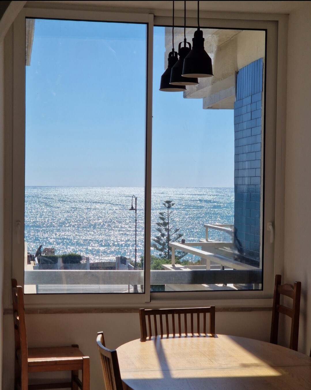 A large window frames a stunning view of the shimmering sea, with sunlight glistening on the water's surface. Below, a wooden table surrounded by chairs is positioned in front of the window, creating an inviting dining area with a coastal vista.