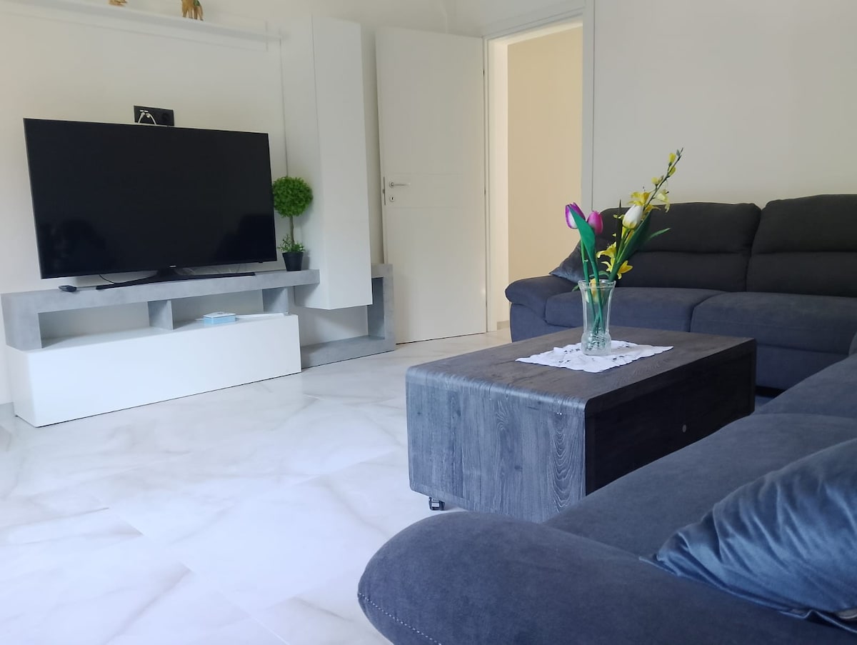 A modern living area features a large, flat-screen television mounted above a minimalist white media console. A dark coffee table is centered on a light marble floor, surrounded by a spacious sectional sofa. A vase with fresh flowers adds a touch of nature to the decor.