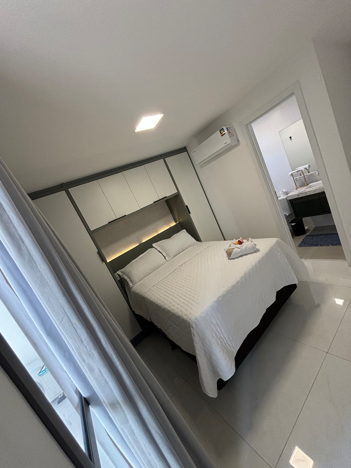 A compact bedroom features a spacious bed with crisp white linens, flanked by subtle lighting elements above. Mirrored cabinetry provides storage, and an air conditioning unit is mounted on the wall. Natural light filters in through a window, reflecting off the glossy tiled floor.