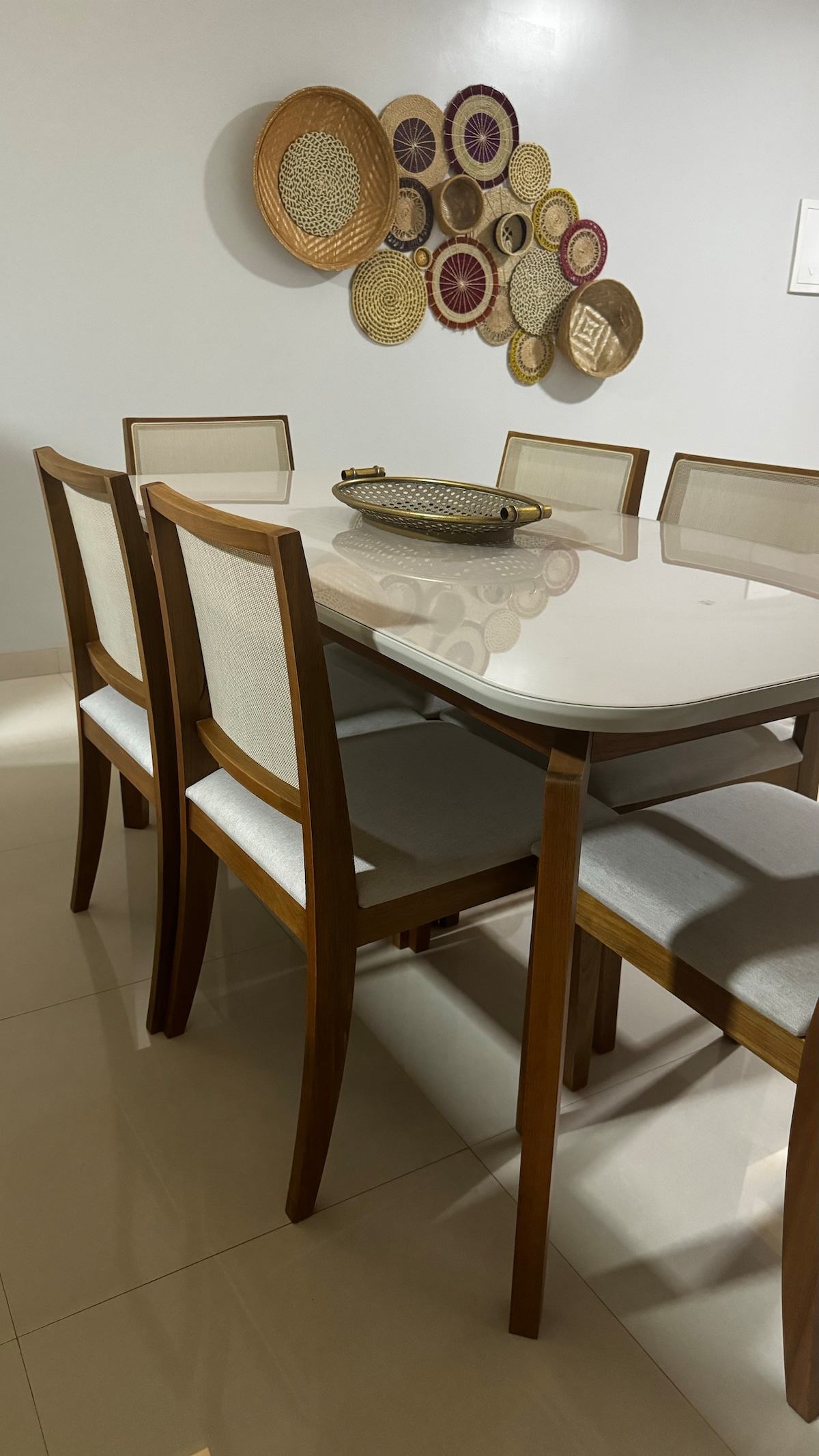 A dining area is showcased with a light-colored table surrounded by six wooden chairs featuring soft seat cushions. Decorative woven wall art in various colors is arranged above the table, adding an artistic touch to the neutral-colored space.