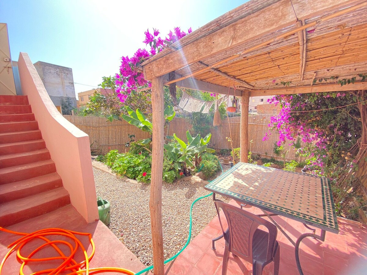 A shaded outdoor seating area features a table surrounded by chairs, with vibrant bougainvillea adding color to the landscape. Lush greenery and gravel paths enhance the garden, while a gently sloping staircase leads to the entrance of the home.