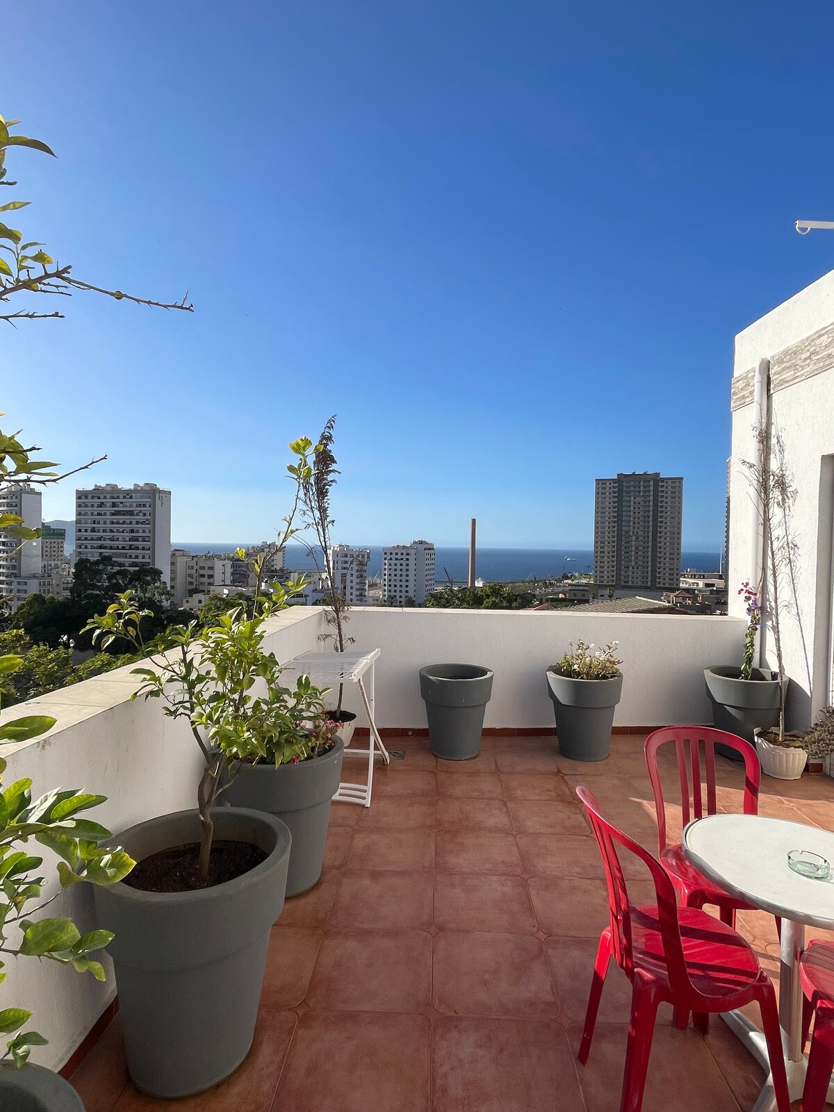 A spacious terrace is shown, adorned with large potted plants and a round table surrounded by red chairs. The view includes a panoramic scene of the city and the sea under a clear blue sky.