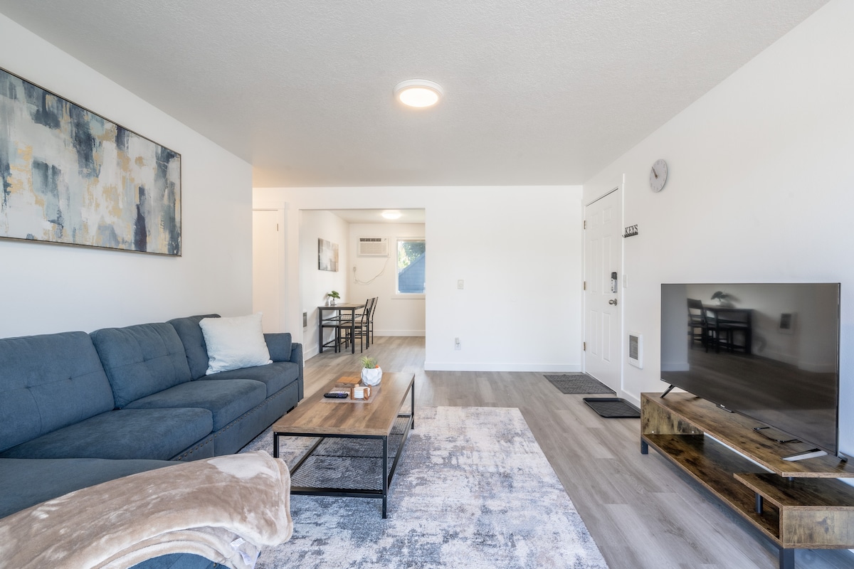 An open living area showcases a comfortable blue sofa and a wooden coffee table on a light area rug. A wall-mounted television is positioned on a modern TV stand. Natural light fills the space from large windows, and a dining area with a table and chairs is visible in the background.