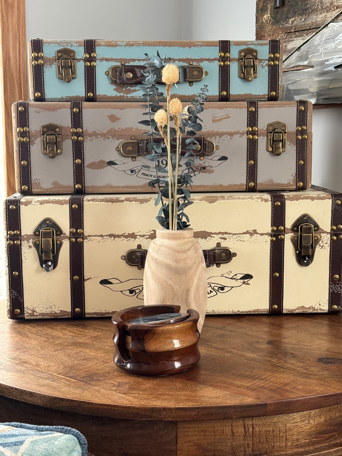 A collection of stacked decorative trunks is displayed on a wooden table, showcasing a variety of muted colors and vintage detailing. A simple wooden vase holds dried flowers, positioned alongside a small, dark wooden bowl, adding a natural element to the arrangement.