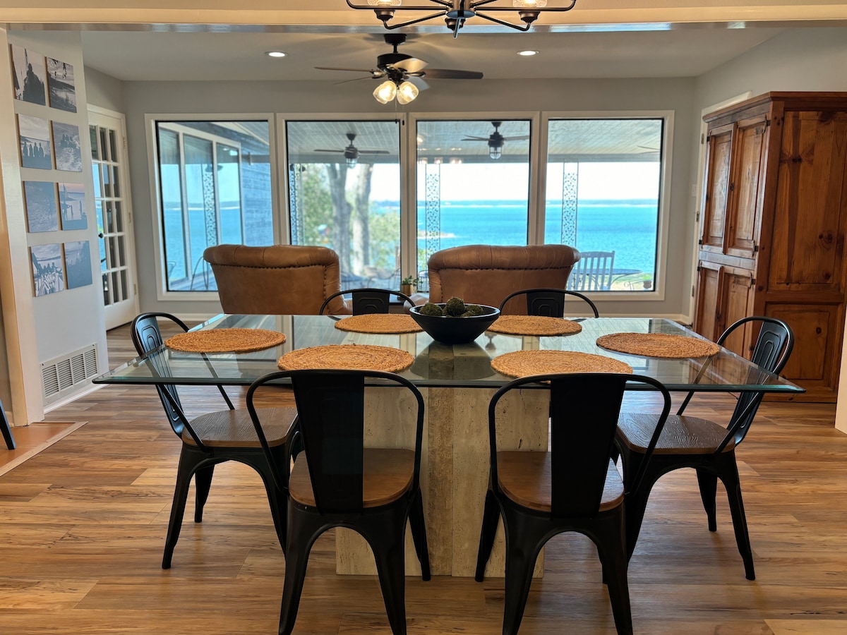A modern dining area is showcased with a clear glass table surrounded by eight chairs. Natural light floods in through large windows offering a view of the lake. Two leather chairs are positioned nearby, enhancing the inviting space for gatherings.