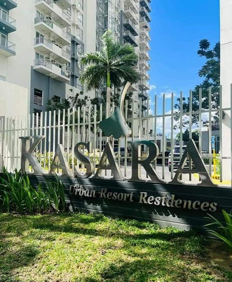 The image displays the entrance sign of Kasara Urban Resort Residences, prominently featured on a well-maintained lawn. Surrounding greenery and a tall palm tree enhance the tropical atmosphere, while modern condominium buildings are visible in the background under a clear blue sky.