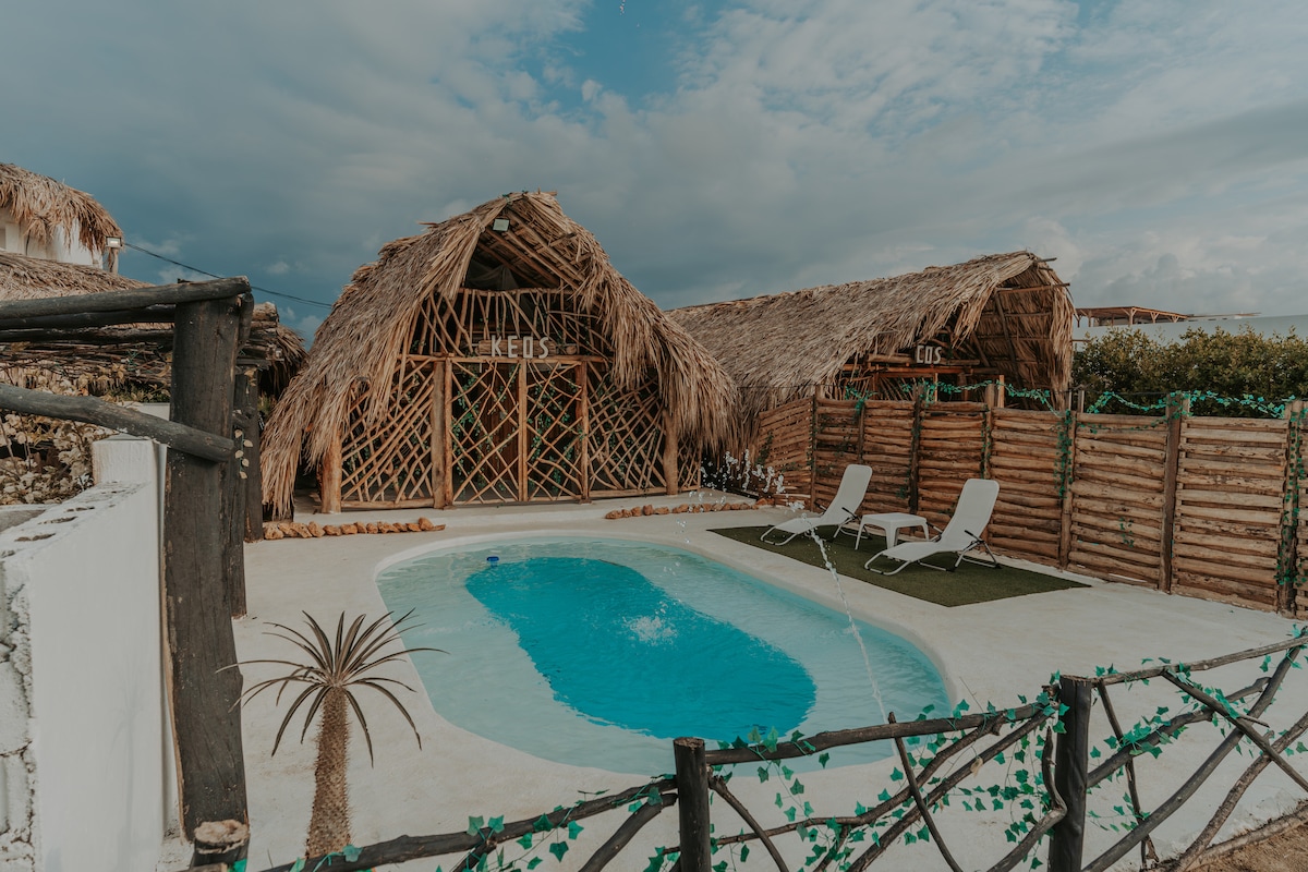 A tranquil outdoor area features a private pool surrounded by a smooth deck. Two comfortable lounge chairs are positioned nearby, and the rustic structures with thatched roofs create a warm atmosphere. Lush greenery lines the space, enhancing the natural setting.