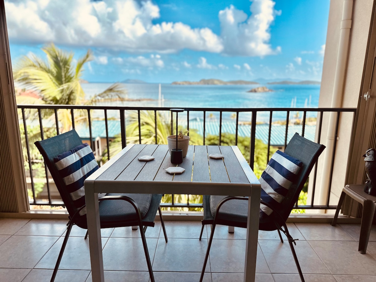A small dining table with two blue and white striped chairs is positioned on a balcony, overlooking the Caribbean Sea. The area is framed by a railing, with the blue water and distant islands in the background, complemented by palm trees swaying gently in the breeze.