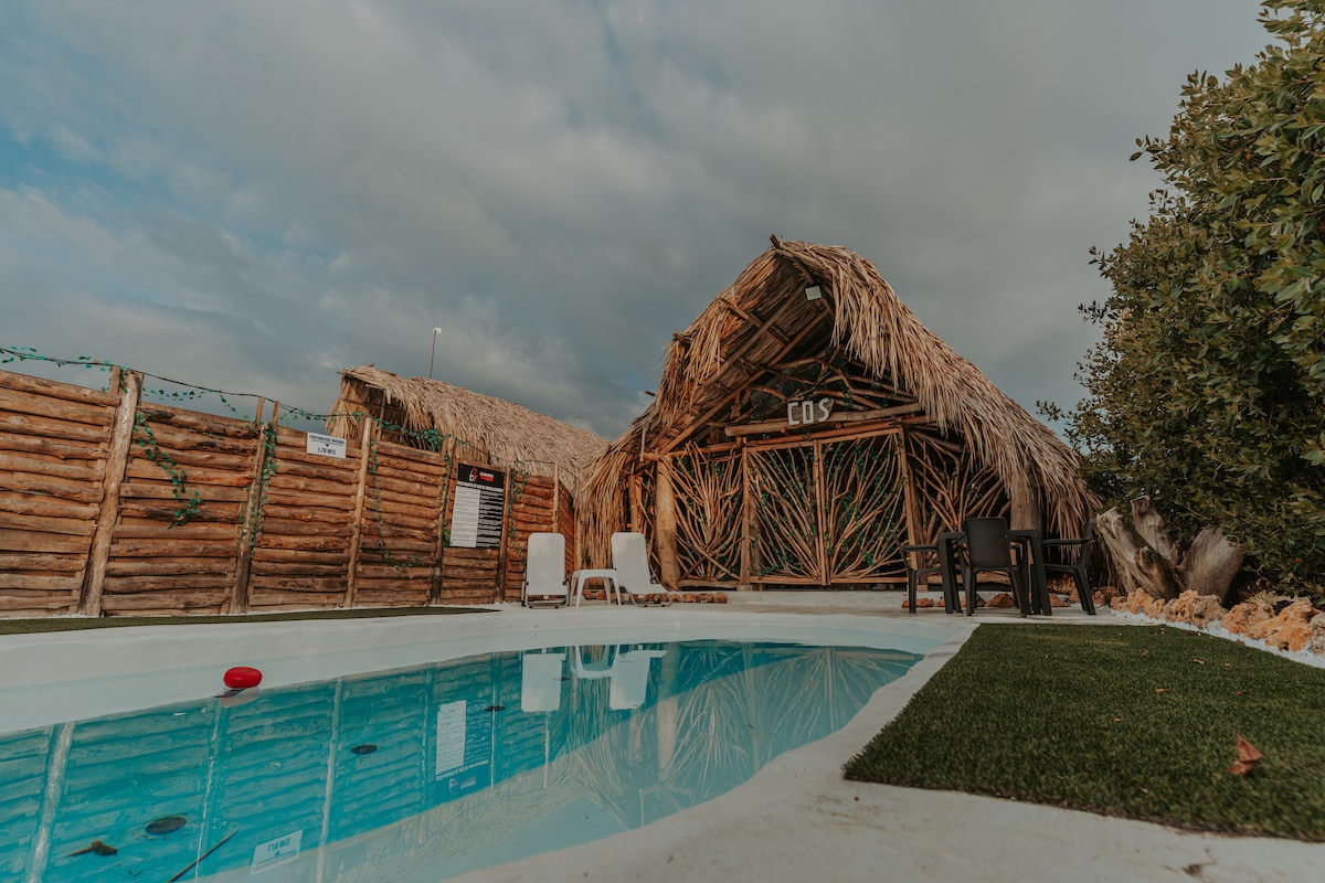 A rustic cabana made of natural materials is seen with a thatched roof, surrounded by wooden fencing. A small swimming pool reflects the cloudy sky, accompanied by white lounge chairs. Green grass lines the pool area, creating a serene outdoor space for relaxation.