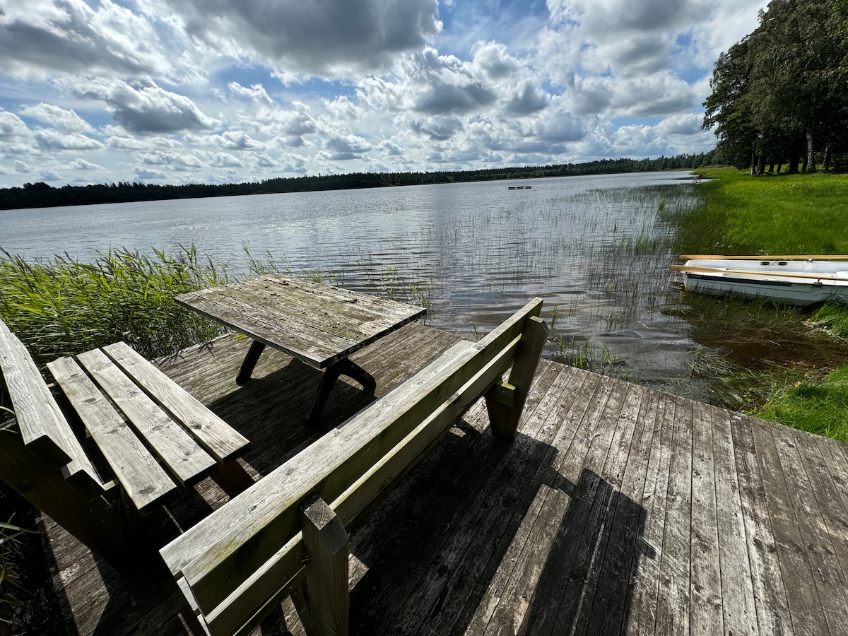 A wooden deck extends over the water's edge, featuring a table surrounded by seating. Tall grasses frame the shoreline, while the serene lake reflects a cloudy sky above. Boats are visible in the distance, adding to the peaceful lakeside setting.