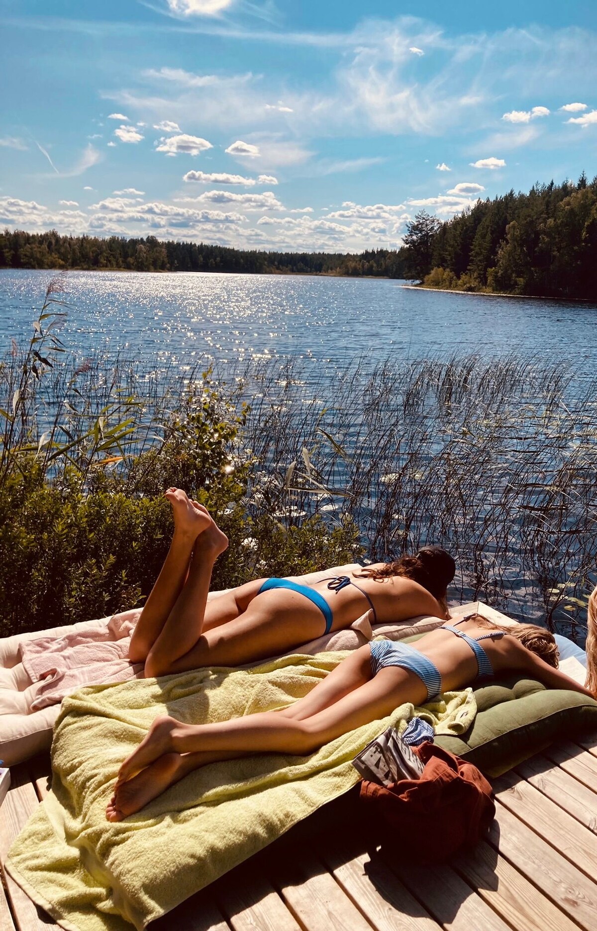 Two individuals are seen resting on a wooden dock by a serene lake, surrounded by lush greenery. Soft blankets are spread out for comfort, while the bright blue sky is dotted with fluffy white clouds. The water reflects the sunlight, enhancing the peaceful setting.