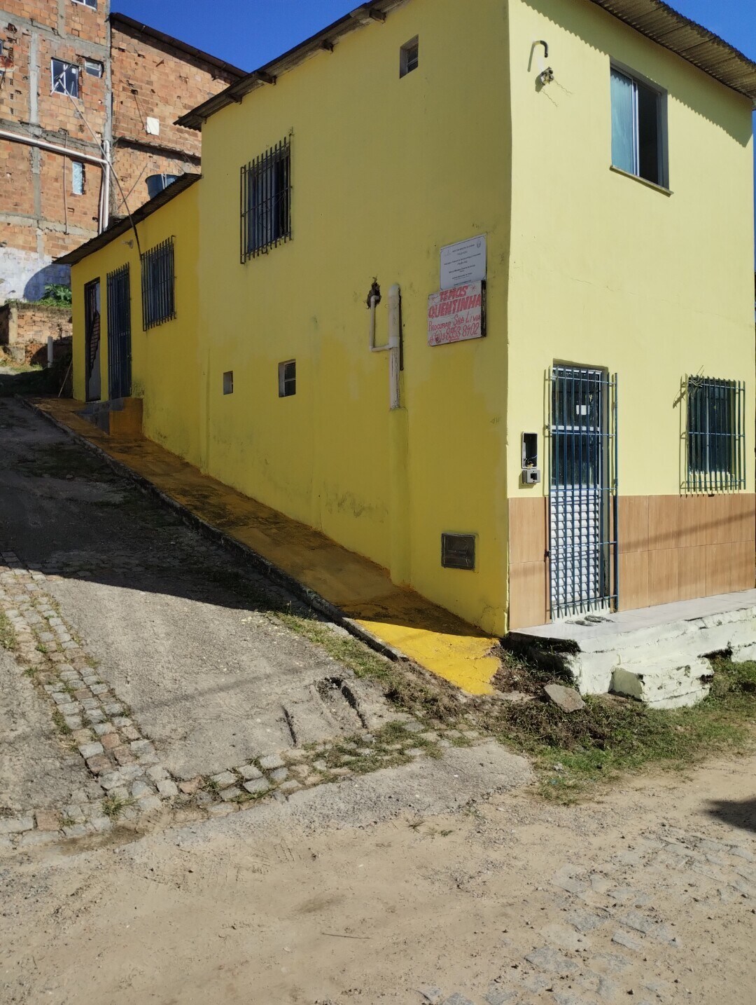 A yellow building is positioned on a sloping pathway, featuring a light-colored exterior with security bars on the windows. A small entrance is visible, highlighting the building's accessibility, while a cobblestone path leads up to the structure.