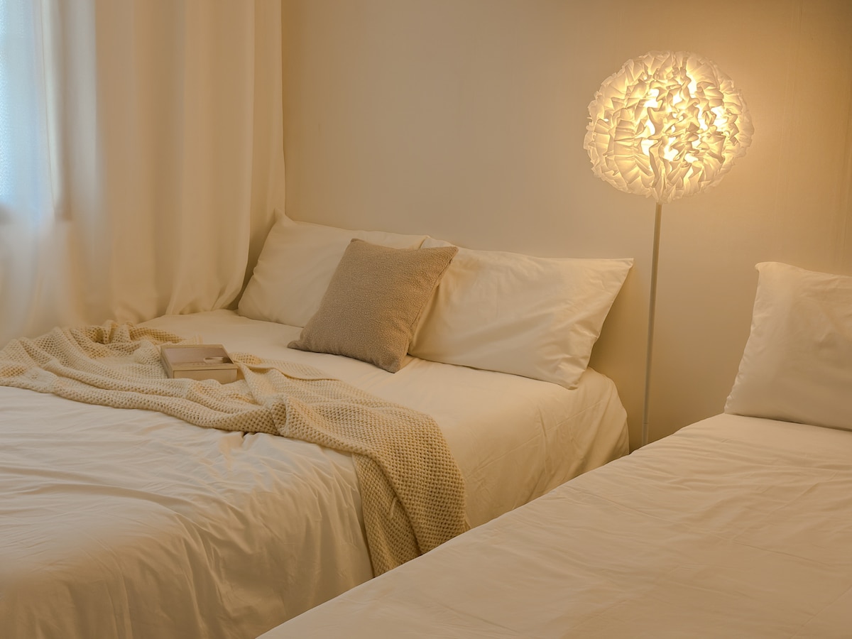 Two cozy beds with white linens are positioned side by side, each adorned with a soft pillow and light throw. A warm, artistic lamp stands on the side, providing gentle lighting. Natural light filters through sheer curtains, creating a serene atmosphere.